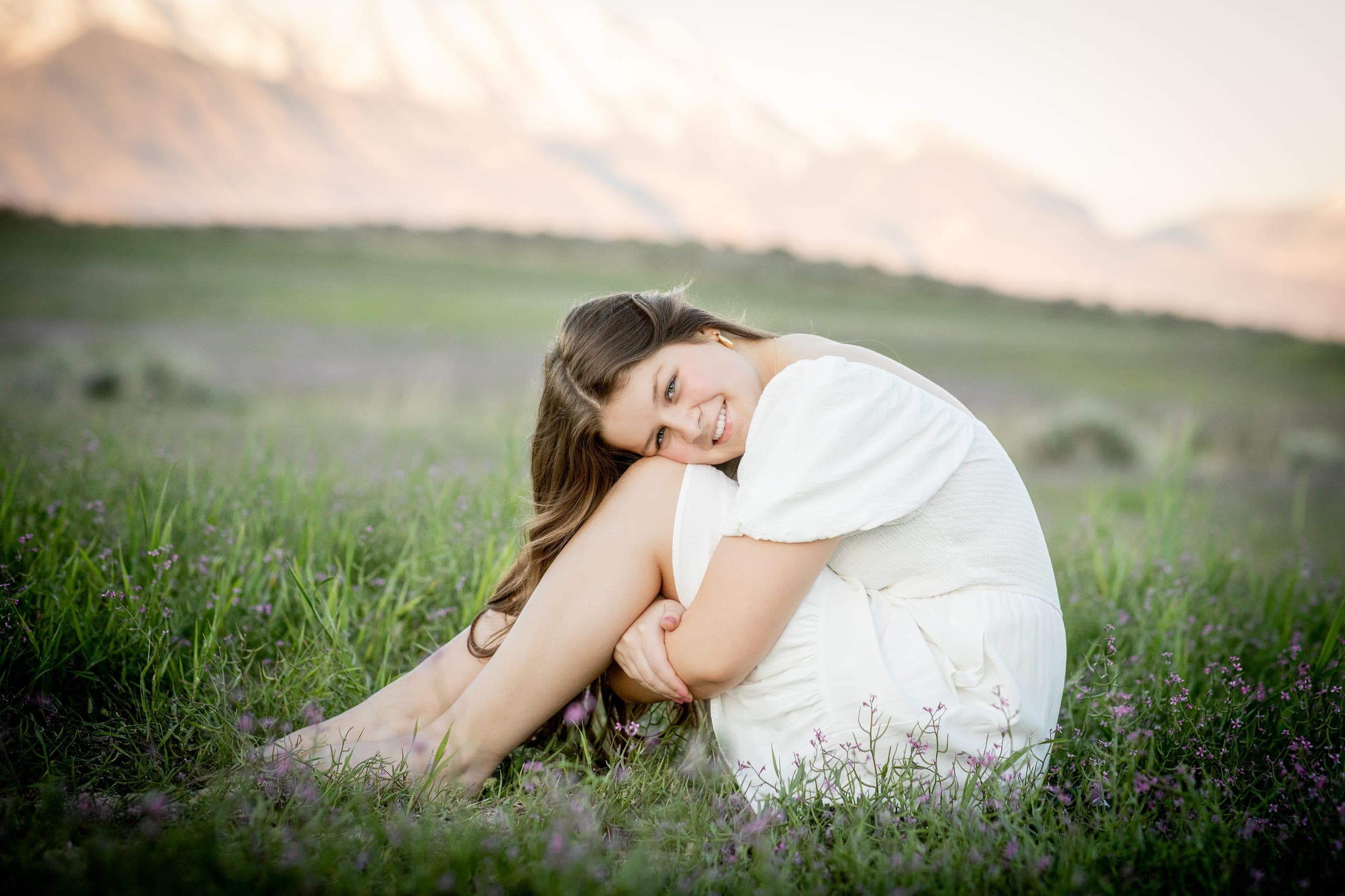 High school senior in a white dress sitting in a grassy field below the mountains