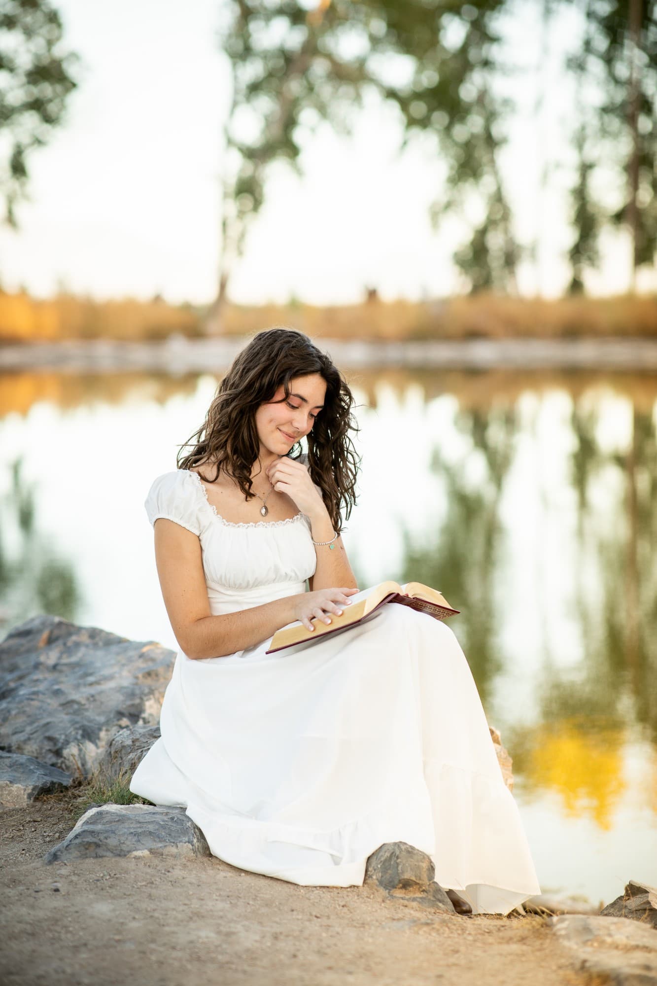 Senior in a white dress reading a book beside a pond