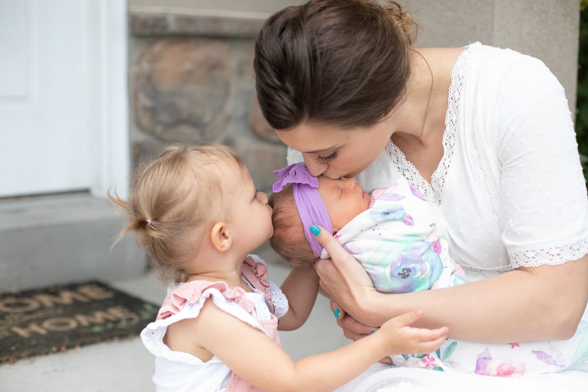 Parent kissing a newborn next to a toddler on the front porch